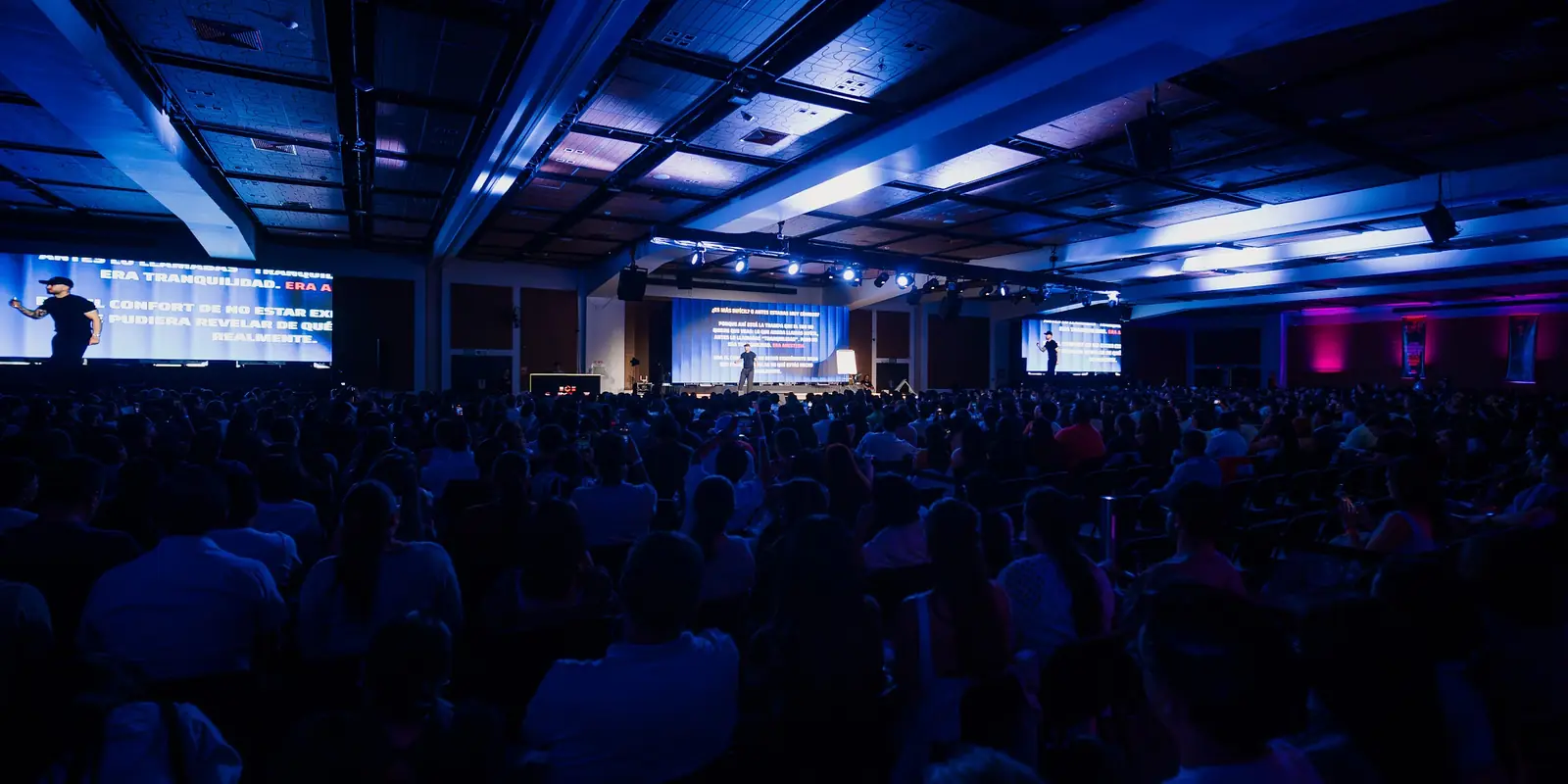 Salón a oscuras lleno de asistentes, con luces azules y pantallas encendidas sobre el escenario.